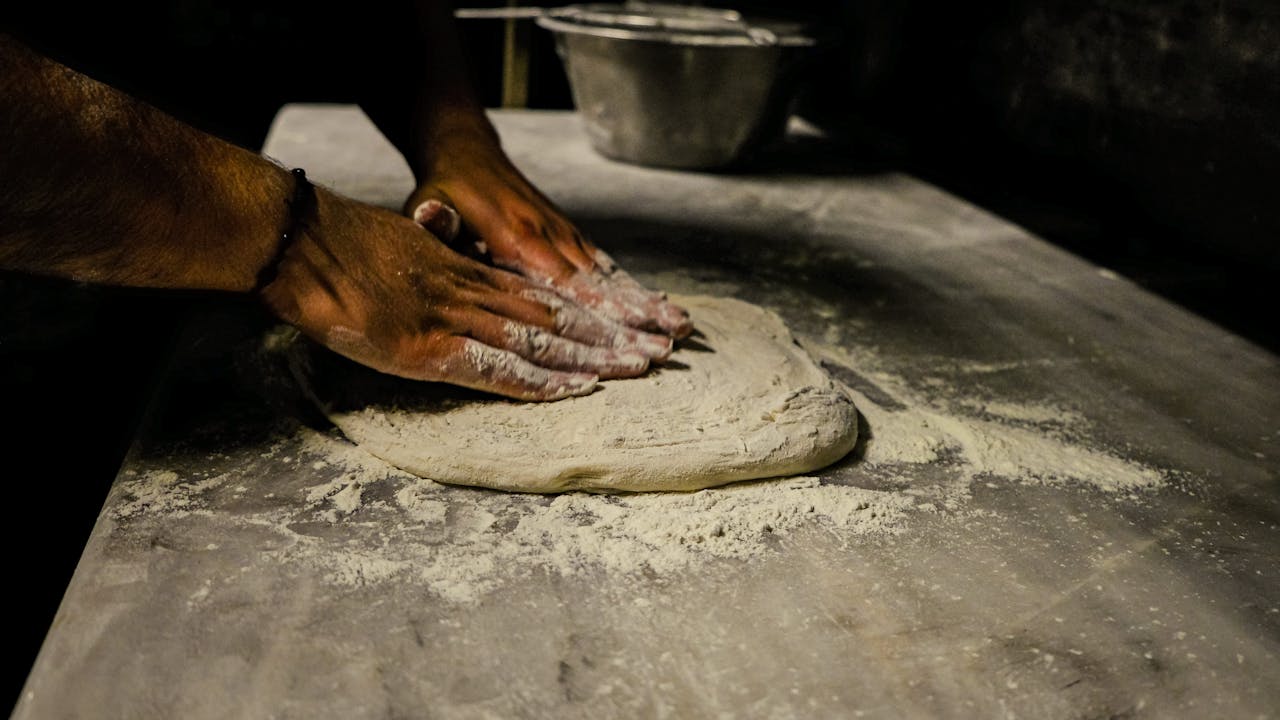 heros-img Close-up of a man kneading dough on a floured table in a traditional setting.
